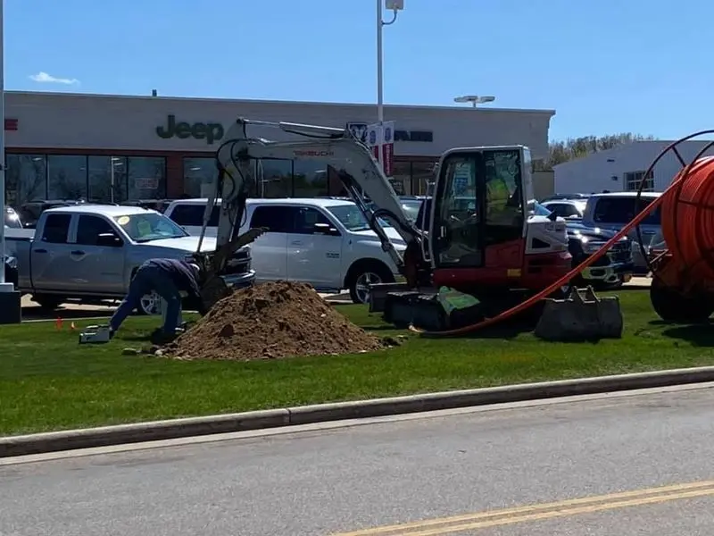 Crew and compact excavator pulling orange conduit into place across a commercial lawn