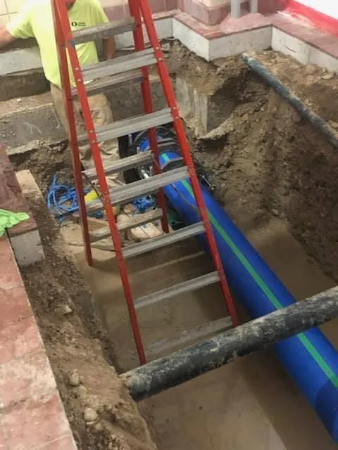 Construction site with ladder in trench; worker in yellow shirt near blue pipe.