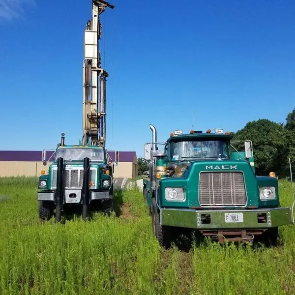 Two green Herr Well Drilling Mack trucks with the drilling mast raised in an open field