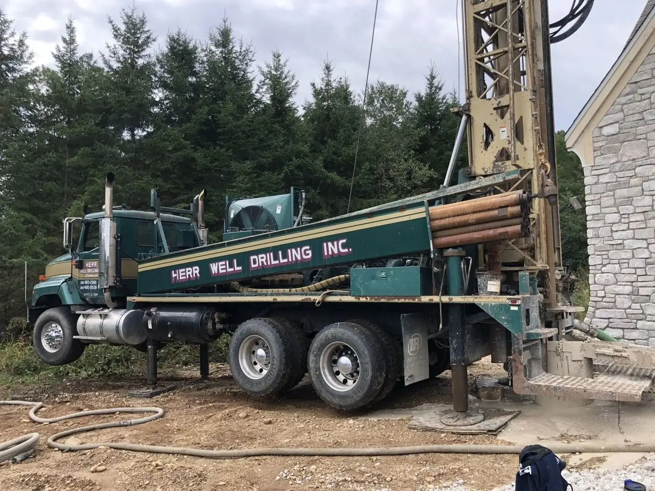 Herr Well Drilling Inc. rig truck with its mast raised at a wooded jobsite beside a stone home.